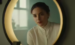 a girl with short hair looking at the camera from behind the mirror