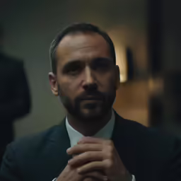 a bearded man looks into the camera with his hands on his chest