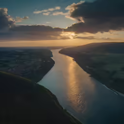 a river next to a hilly green hillside with sun behind it