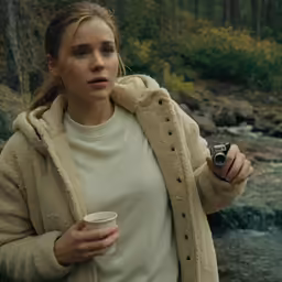 woman in coat holding coffee cup and camera near stream
