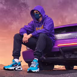 the man sits on the hoodie and wears sneakers,a man wearing a purple hoodie and nike sneakers sits on the front of a sports car