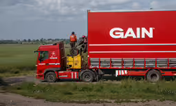 a man stands in front of a giant red container truck,a red semi trailer is parked next to a rural field