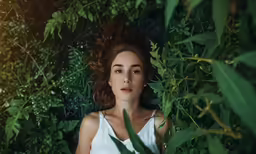 a woman in a white top sitting by a bush