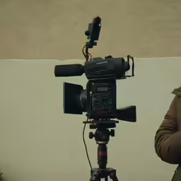 a man stands near a camera with his arm resting on the tripod