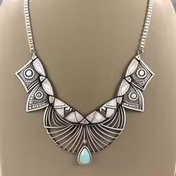 the silver and turquoise necklace is on display,a very pretty white and silver necklace on display,an intricately designed necklace with tear shaped turquoise stone,a silver necklace that is on a mannequin,a silver necklace with a tear shaped drop