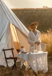 a lady that is standing up in front of a tent,a woman in white shirt standing next to a table with a tea set up