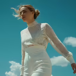 a woman standing against a blue sky and clouds wearing a sheer white top
