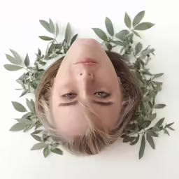 a woman wearing a black headband and eyes closed looks up with eyes wide open, surrounded by green leaves