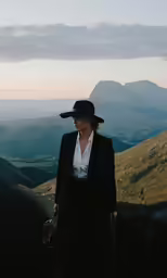 a woman wearing a suit and hat while standing on a mountain