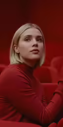 a woman sitting in an empty auditorium wearing a red turtle neck
