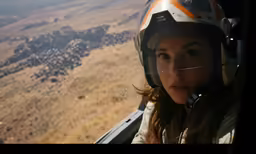 a girl is in the cockpit of a airplane looking down at an open plain with a mountain in the background