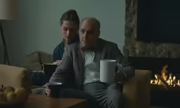 a man and woman sit near a fireplace with cups in their hands