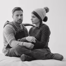 a young man and woman in winter clothes sit on a bed, looking at each other and smiling