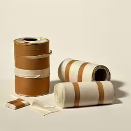 a roll of white toilet paper next to a roll of brown striped toilet paper,three different types of material, one white and one brown,several different rolls of yarn are laying on a white surface,an orange and white stripe is next to a roll of yarn,two rolls of twine are laid out on a surface