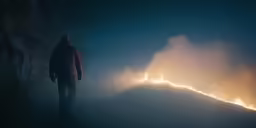 a man in a jacket standing next to a mountain that is in flames