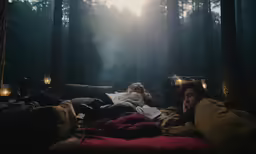 two people in the woods at night lying on a bed with pillows