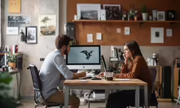 two people sit at a desk and look at a computer