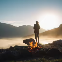 a woman stands on a rock in the foreground, near an open fire and fog