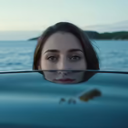 the girl looks through a windshield as the water and sky reflect on the vehicle