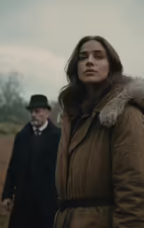 two people in long coats stand in an open field