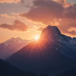the sun setting behind a tall mountain with a cloud filled sky