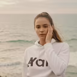 a girl is holding her face with one hand and standing in front of the ocean