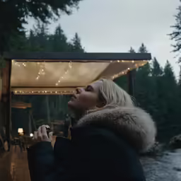 a blonde woman dressed in winter clothes holds a small glass