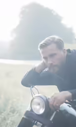 man with beard and black jacket leaning on motorcycle