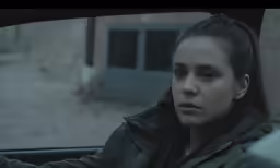 a woman sits in the passenger seat of a car and looks into the camera