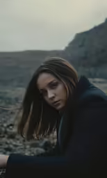 a woman in a jacket sitting on rocks and staring into the distance