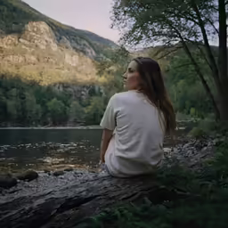a woman sitting on the shore of a lake by trees
