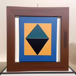 an abstract painting in brown frames with blue and yellow shapes,an abstract painting hanging in the middle of a wall,an art piece sits in front of a wall,the painting is on a table and the wall,an abstract piece hanging on the wall in a room