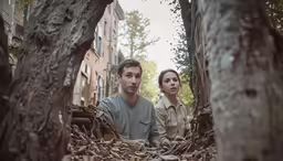 two people standing in the woods with a pile of leaves on it