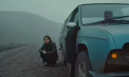 a man sitting beside a blue car on a gravel road