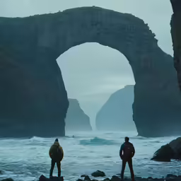 two men with their backs turned to the camera look out at an arch bridge on the ocean