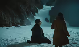the woman sits in front of a waterfall and a woman in a long black coat sits behind