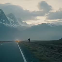 a man standing at the front of a vehicle while two others drive down a road in the evening
