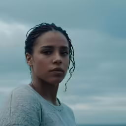 young woman with black curly hair standing in front of an ocean