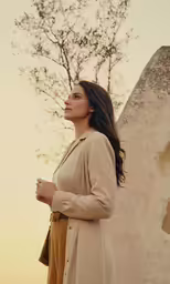 woman in trench coat standing near wall at sunset