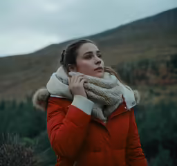 a beautiful woman standing in the mountains holding her scarf around her neck
