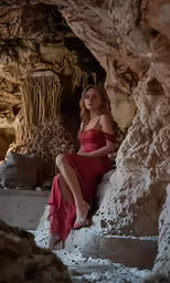a woman sitting in a cave wearing a red dress