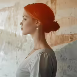 a woman in white with red hair standing with her head tilted and looking away