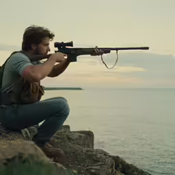 man sitting on rock with rifle looking through scope