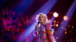 a woman wearing a mask and holding a microphone,the singer is wearing feathers and singing into the microphone