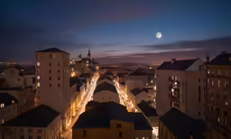 a view of a city at night with lights all over the buildings and moon overhead
