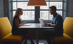 a man and a woman are sitting at a table in front of a laptop