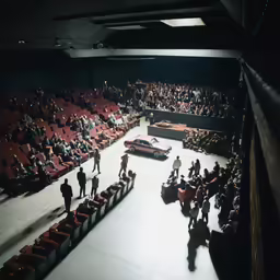 a large theatre with rows of seats and a stage in the center of it, with two rows of seats on each side, all facing a small auditorium that is full, with several people looking at a car