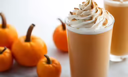 two glasses of a latte with whipped cream and pumpkins behind them
