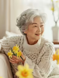 an older asian woman smiles and holds some daffodils,a woman sitting on a couch holding flowers in her hands