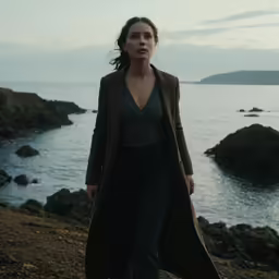 a woman walking along the water with a dark coat on